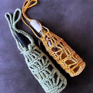 Könnte beinhalten: Zwei gehäkelte Flaschenhalter mit langen Trageriemen. Einer ist salbeigrün und enthält eine dunkle Glasflasche, während der andere goldgelb ist und eine klare Glasflasche mit weißem Verschluss hält. Beide haben ein offenes Häkelmuster.