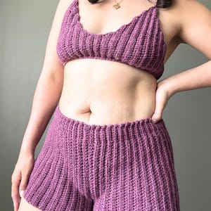 May include: A two-piece set of handmade crochet clothing in a deep purple hue. The set includes a bra-style top with thin straps and high-waisted shorts. The ribbed texture of the crochet adds visual interest to the garments.