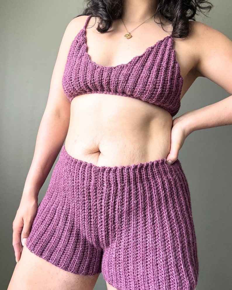 May include: A two-piece set of handmade crochet clothing in a deep purple hue. The set includes a bra-style top with thin straps and high-waisted shorts. The ribbed texture of the crochet adds visual interest to the garments.