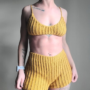 May include: Mustard yellow crochet bikini top and matching shorts set. The top has thin straps and a ribbed texture. The shorts are high-waisted with a similar ribbed design. The set is handmade and perfect for summer.