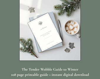 May include: A white document titled The Tender Wobble Guide to Winter, A Kinder Way to Move Through The Colder Months, resting on a light blue notebook. A silver pen, a mug with marshmallows, and green pine sprigs are arranged around the document.
