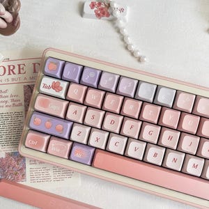 Puede incluir: Un teclado mecánico de colores pastel con teclas rosas, lavanda y blancas. El teclado tiene un marco rosa y una barra espaciadora a juego. Algunas teclas presentan diseños de frutas y flores. La imagen también incluye algunos elementos decorativos.