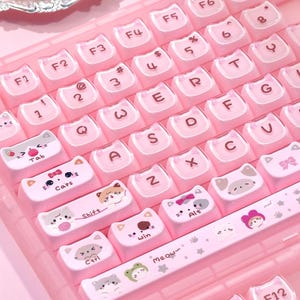 May include: A pink keyboard with white keycaps featuring cat faces and other cute illustrations. The keyboard has a standard QWERTY layout with function keys and a numeric keypad. The keycaps have a light pink background with white lettering.