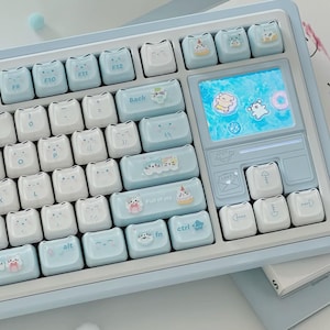 May include: A light blue and white mechanical keyboard featuring custom cat-themed keycaps with various facial expressions and small illustrations. The keyboard includes a rectangular digital display screen showing animated characters on a blue background.