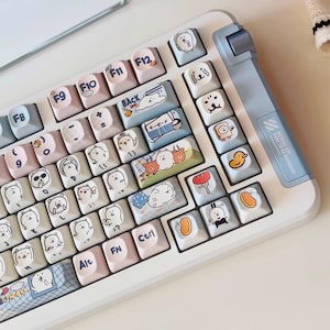 May include: A white mechanical keyboard with custom keycaps featuring cartoon animal illustrations. The keycaps are a mix of pastel colors and include text and graphics. Additional keycaps and accessories are scattered around the keyboard.