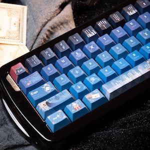 May include: A custom mechanical keyboard with blue keycaps featuring various illustrations and symbols. The keycaps have a gradient effect, transitioning from dark blue to light blue. The keyboard has a dark wooden frame.