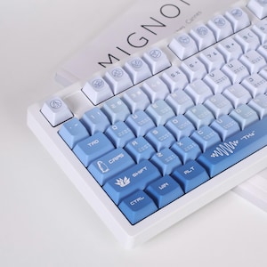 May include: A white mechanical keyboard with blue gradient keycaps. The keycaps transition from white to light blue to a deep blue. Some keys have unique icons. The word "MIGNON" is visible in the background.