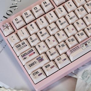 Könnte beinhalten: Nahaufnahme einer pinkfarbenen mechanischen Tastatur mit cremefarbenen Tastenkappen. Die Tastenkappen haben ein Rastermuster und verschiedene Symbole und Texte, darunter "TAB", "SHIFT" und "CTRL". Die Tastatur hat einen hellrosa Rahmen.