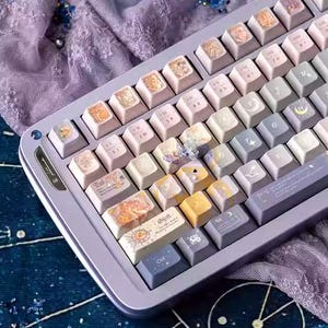 Op de afbeelding: Een aangepast mechanisch toetsenbord met een lichtpaars frame en keycaps in verschillende pastelkleuren. De keycaps hebben unieke ontwerpen en symbolen, met een paar gele en blauwe toetsen. Het toetsenbord ligt op een donkerblauwe ondergrond.
