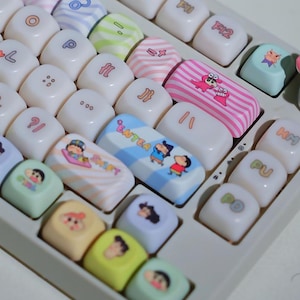 May include: A close-up shot of a mechanical keyboard with custom keycaps. The keycaps display colourful designs, including cartoon characters, stripes, and symbols. The keyboard has a light grey base and white keycaps with the text "ENTER" on one key.