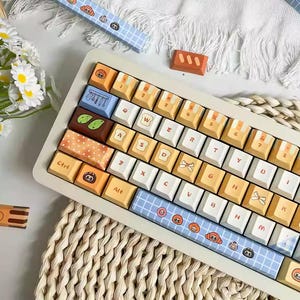 May include: A custom mechanical keyboard with a cream-colored frame and colorful keycaps. The keycaps feature various designs, including food-themed illustrations and patterned accents. The keyboard also has blue accent bars with cartoon graphics.