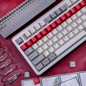 May include: A mechanical keyboard with white, red, and grey keycaps. The keyboard has a white frame and a variety of keycap designs. A calendar showing April 2025 is in the background. Other accessories are also visible.