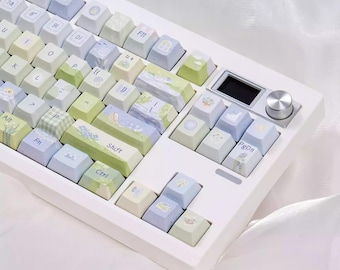 Könnte beinhalten: Eine weiße mechanische Tastatur mit pastellfarbenen Tastenkappen. Die Tastenkappen weisen verschiedene Designs auf, darunter Blumenmuster und Landschaftsszenen. Die Tastatur hat einen kleinen Bildschirm und einen silbernen Knopf auf der rechten Seite.