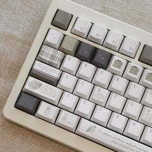 Può includere: Una tastiera meccanica beige con tasti personalizzati. I tasti presentano un mix di bianco, grigio e motivi, tra cui illustrazioni botaniche e testo. La barra spaziatrice ha il testo "In rainforest expeditions, medicinal compounds are plants-potential cures hidden in foliage."