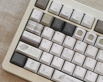 May include: A beige mechanical keyboard with custom keycaps. The keycaps feature a mix of white, gray, and patterned designs, including botanical illustrations and text. The space bar has text that reads "In rainforest expeditions, medicinal compounds are plants-potential cures hidden in foliage."