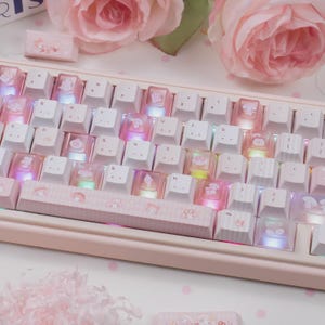 May include: A pink and white mechanical keyboard with custom keycaps. The keycaps feature cartoon characters and are translucent, allowing the colorful LED backlighting to shine through. The keyboard has a matching pink frame.