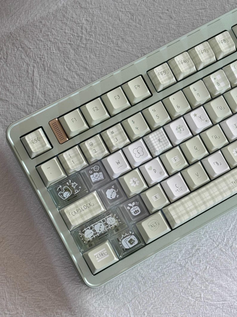 Puede incluir: Un teclado mec&aacute;nico verde menta con teclas blancas y estampadas. Algunas teclas tienen cubiertas transparentes con ilustraciones de dibujos animados. El teclado tiene un marco met&aacute;lico y una variedad de teclas de funci&oacute;n.