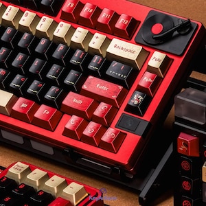 Op de afbeelding: Een mechanisch toetsenbord met een rood frame, een mix van zwarte, rode en gouden toetsen, en een draaiknop in de rechterbovenhoek. De toetsen bevatten aangepaste letters, waaronder 'Backspace', 'Enter', 'Shift', 'PgUp', 'PgDn' en 'Fn'.