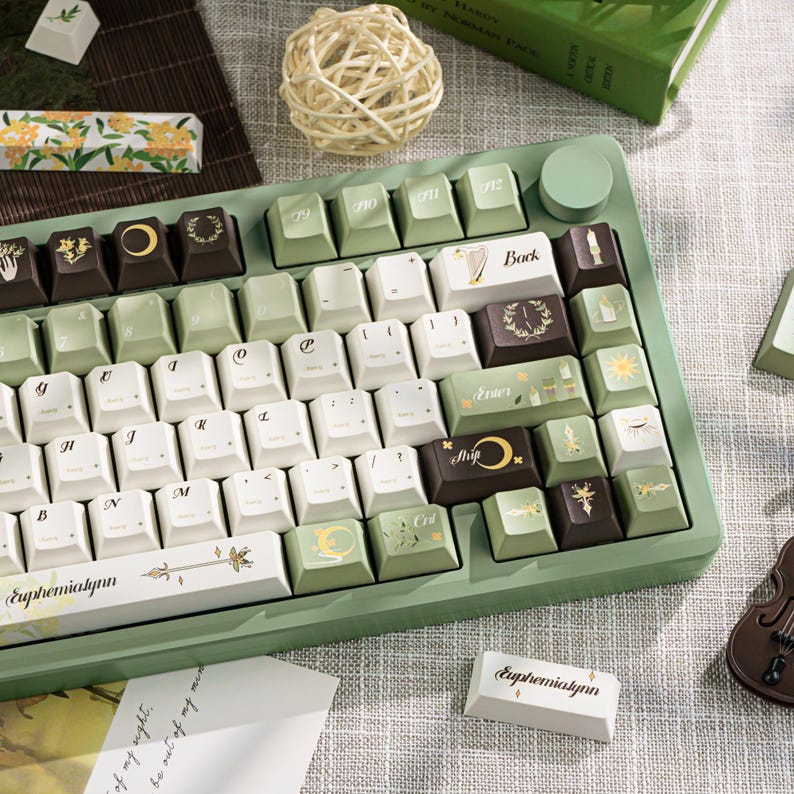 Puede incluir: Un teclado mec&aacute;nico personalizado con una base verde claro y teclas blancas, verdes y marrones. Las teclas presentan varios s&iacute;mbolos y dise&ntilde;os, incluyendo lunas, soles y patrones florales. El teclado tiene el texto "Euphemialynn" en una tecla.