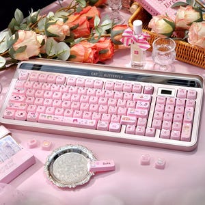 May include: A pink keyboard with cat-themed keycaps and the text "CAT BUTTERFLY" is displayed on a pink surface. The keyboard is surrounded by decorative items, including flowers, a mirror, and a perfume bottle. The overall aesthetic is soft and feminine.