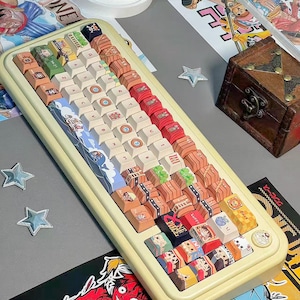 May include: A custom keyboard with a cream-colored frame and a variety of colorful keycaps. The keycaps feature illustrations of anime characters and other designs. The keyboard is surrounded by related artwork and a small wooden chest.