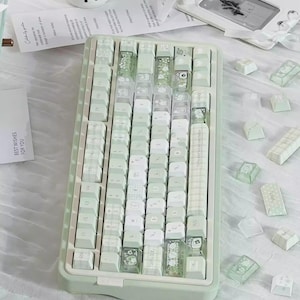 Puede incluir: Un teclado mec&aacute;nico verde menta con teclas blancas y transparentes. Algunas teclas tienen dise&ntilde;os decorativos. El teclado est&aacute; rodeado de teclas adicionales y una etiqueta blanca con el texto "Rosy Posy+".