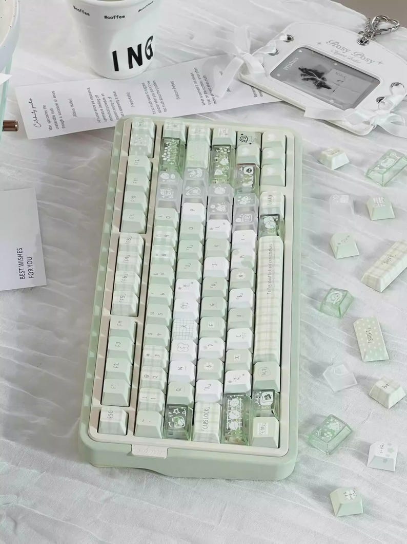 Puede incluir: Un teclado mec&aacute;nico verde menta con teclas blancas y transparentes. Algunas teclas tienen dise&ntilde;os decorativos. El teclado est&aacute; rodeado de teclas adicionales y una etiqueta blanca con el texto "Rosy Posy+".