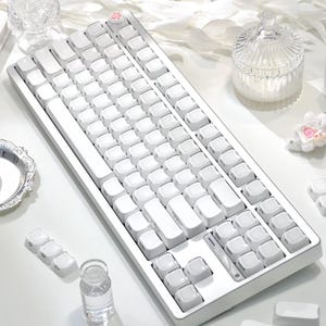 May include: A white mechanical keyboard with white keycaps. The keyboard has a minimalist design with a pink paw print key. Additional white keycaps are scattered around the keyboard.
