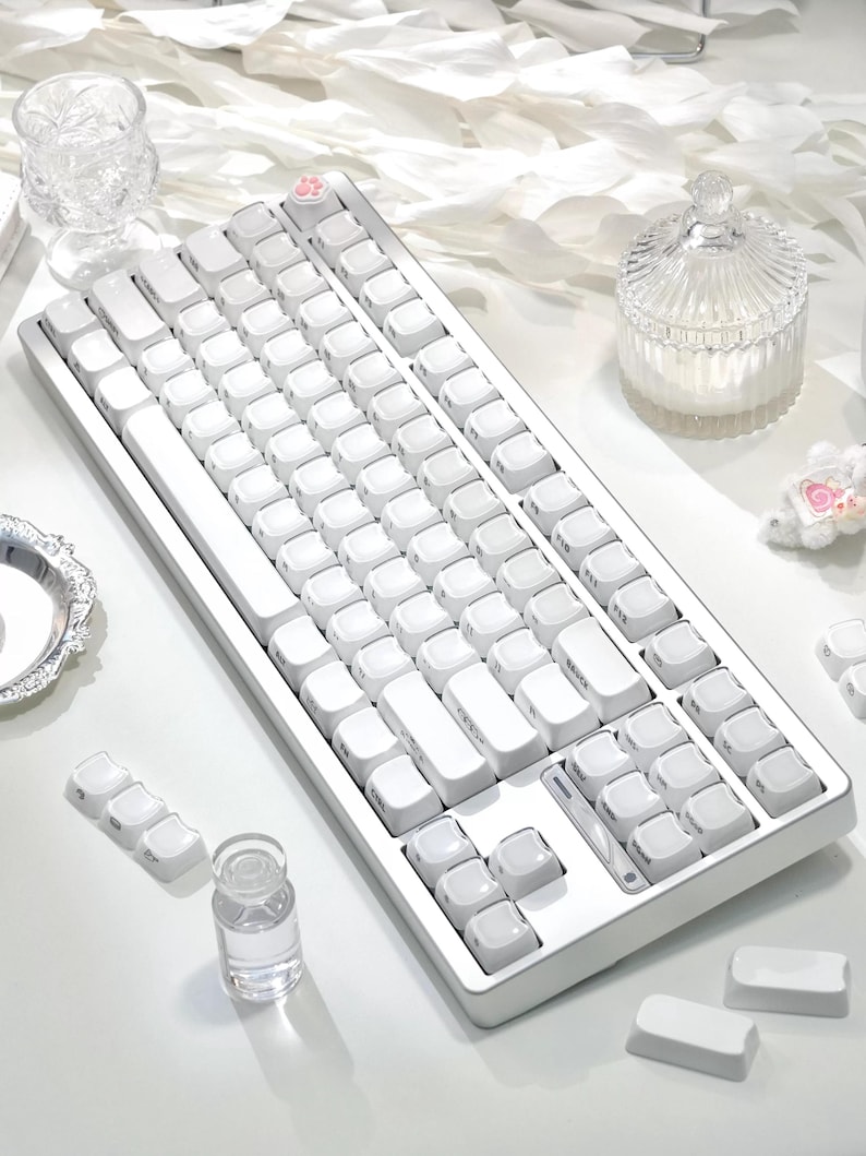 May include: A white mechanical keyboard with white keycaps. The keyboard has a minimalist design with a pink paw print key. Additional white keycaps are scattered around the keyboard.