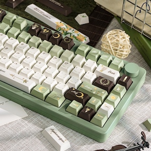 Puede incluir: Un teclado mec&aacute;nico verde y blanco con teclas personalizadas. Las teclas presentan varios dise&ntilde;os, incluyendo patrones florales, lunas y soles. El teclado tiene una base verde y una perilla de control redonda.