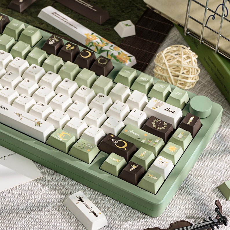 Puede incluir: Un teclado mec&aacute;nico verde y blanco con teclas personalizadas. Las teclas presentan varios dise&ntilde;os, incluyendo patrones florales, lunas y soles. El teclado tiene una base verde y una perilla de control redonda.