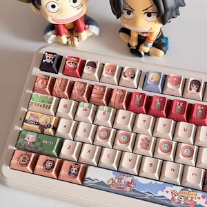 May include: A custom mechanical keyboard with keycaps featuring anime characters and imagery. The keyboard has a white frame with various colorful keycaps. Two figurines of anime characters are placed above the keyboard.