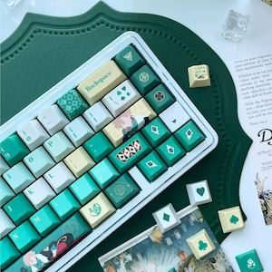Può includere: Una tastiera meccanica con una cornice bianca e una varietà di tasti bianchi, verde acqua e verdi. Alcuni tasti presentano simboli di carte da gioco e disegni decorativi. La tastiera è su una superficie verde.