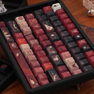May include: A selection of custom keyboard keycaps presented in a black tray. The keycaps are predominantly burgundy, black, and cream. Several keycaps display illustrations of flowers, butterflies, and other decorative elements.