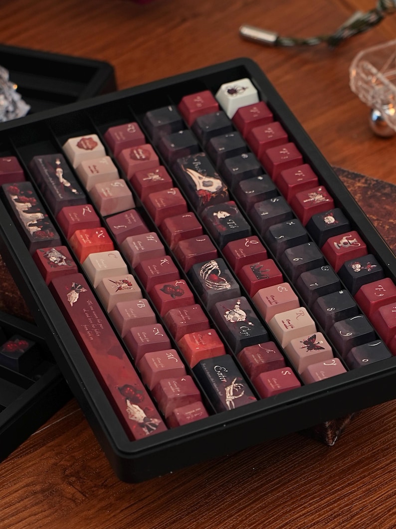 May include: A selection of custom keyboard keycaps presented in a black tray. The keycaps are predominantly burgundy, black, and cream. Several keycaps display illustrations of flowers, butterflies, and other decorative elements.