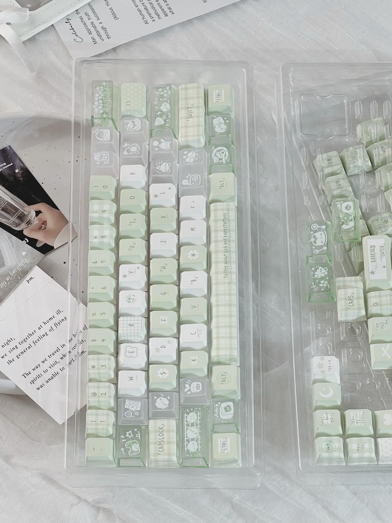 Puede incluir: Un conjunto de teclas de teclado verde claro y blanco en un recipiente de pl&aacute;stico transparente. Las teclas presentan varios dise&ntilde;os, incluyendo cuadros, florales y dibujos animados. Algunas teclas tienen texto como "CAPSLOCK" y "CTRL".