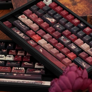 May include: A selection of custom keyboard keycaps displayed in a black tray. The keycaps feature various designs in red, brown, and cream hues, with some displaying illustrations and text. The keycaps are arranged in rows, highlighting their unique artwork.