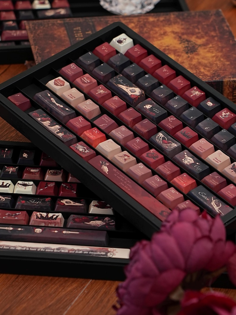 May include: A selection of custom keyboard keycaps displayed in a black tray. The keycaps feature various designs in red, brown, and cream hues, with some displaying illustrations and text. The keycaps are arranged in rows, highlighting their unique artwork.