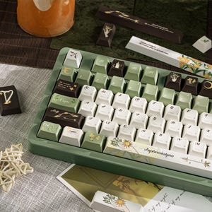 Puede incluir: Un teclado mec&aacute;nico personalizado con teclas blancas, verdes y marrones. Las teclas presentan dise&ntilde;os bot&aacute;nicos y celestiales, con la palabra "Euphemialynn" impresa en la barra espaciadora. El teclado tiene un marco verde.