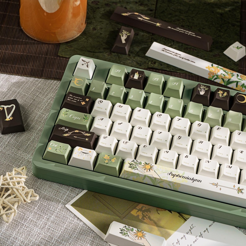 Puede incluir: Un teclado mec&aacute;nico personalizado con teclas blancas, verdes y marrones. Las teclas presentan dise&ntilde;os bot&aacute;nicos y celestiales, con la palabra "Euphemialynn" impresa en la barra espaciadora. El teclado tiene un marco verde.