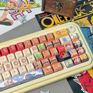 May include: A custom keyboard with keycaps featuring characters and imagery from the anime series One Piece. The keyboard has a cream-colored frame and a variety of colorful keycaps with unique designs. A small wooden treasure chest sits above the keyboard.