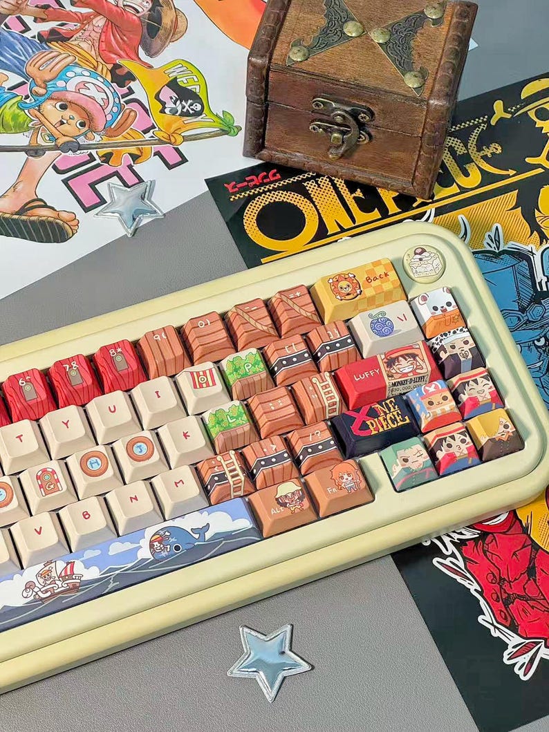 May include: A custom keyboard with keycaps featuring characters and imagery from the anime series One Piece. The keyboard has a cream-colored frame and a variety of colorful keycaps with unique designs. A small wooden treasure chest sits above the keyboard.