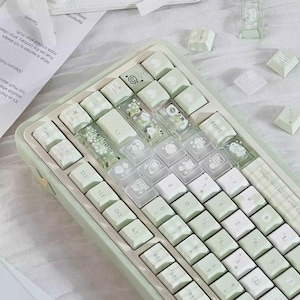 Puede incluir: Un teclado mec&aacute;nico verde menta con teclas blancas. Algunas teclas son transparentes con elementos decorativos. El teclado tiene un dise&ntilde;o compacto e incluye teclas de funci&oacute;n. Teclas adicionales y un peque&ntilde;o art&iacute;culo decorativo transparente est&aacute;n dispersos.