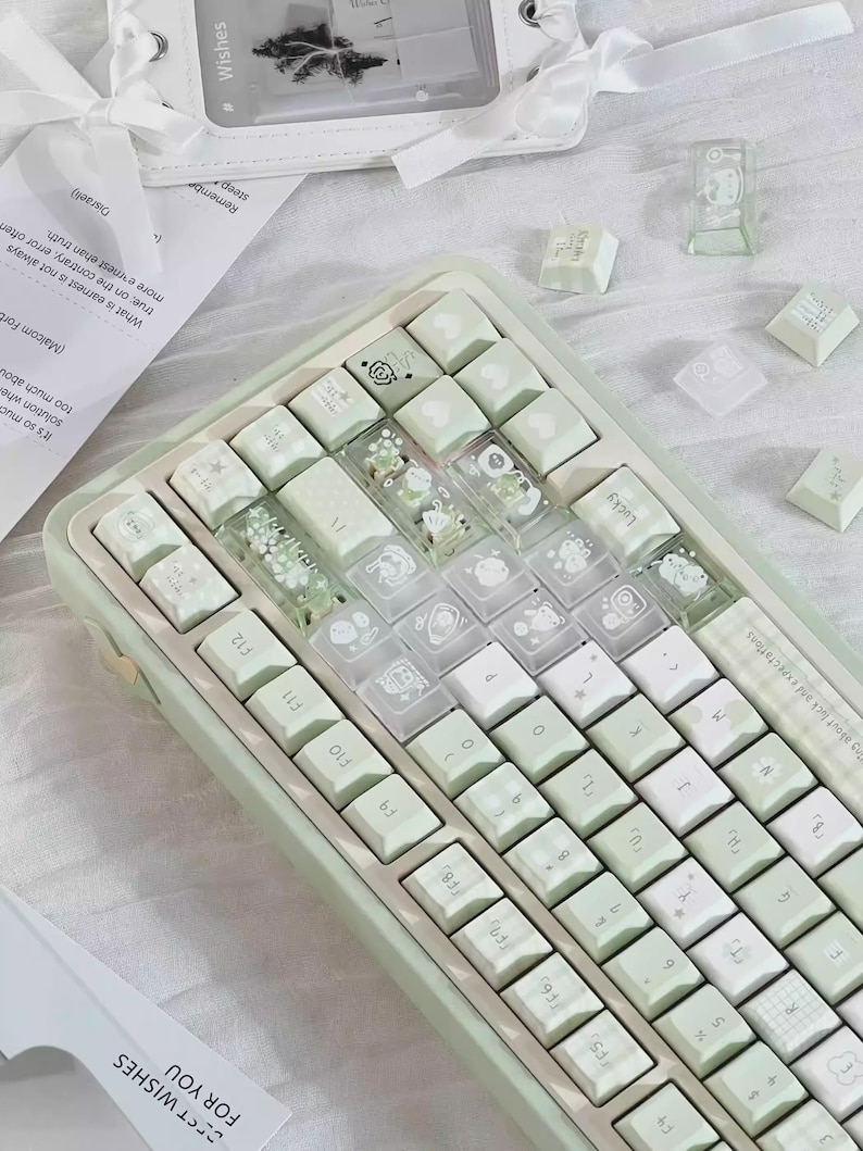 Puede incluir: Un teclado mec&aacute;nico verde menta con teclas blancas. Algunas teclas son transparentes con elementos decorativos. El teclado tiene un dise&ntilde;o compacto e incluye teclas de funci&oacute;n. Teclas adicionales y un peque&ntilde;o art&iacute;culo decorativo transparente est&aacute;n dispersos.