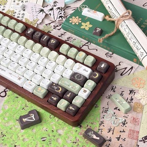 Puede incluir: Un teclado mec&aacute;nico personalizado con marco de madera y teclas en tonos blanco, verde y marr&oacute;n. Las teclas presentan varios s&iacute;mbolos y dise&ntilde;os. Teclas adicionales y elementos decorativos, incluyendo un pergamino y papel con caligraf&iacute;a, est&aacute;n dispuestos alrededor del teclado.