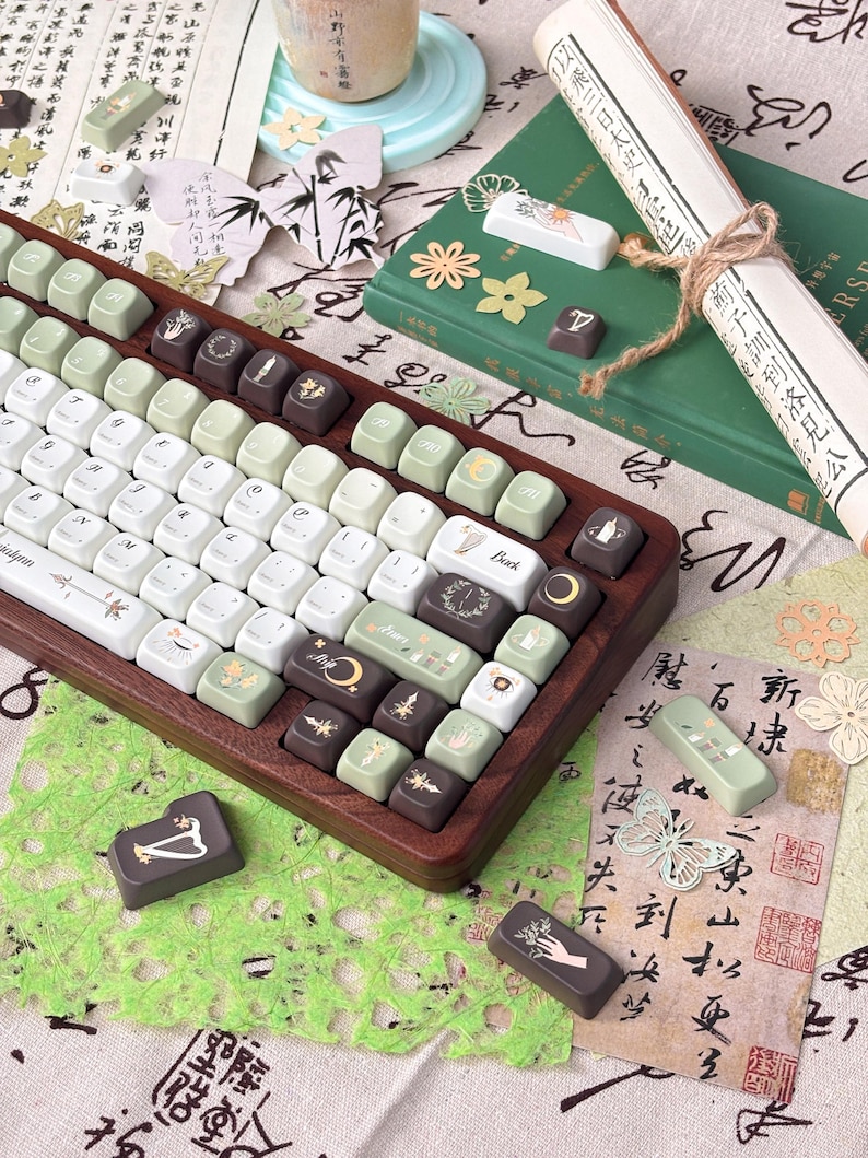 Puede incluir: Un teclado mec&aacute;nico personalizado con marco de madera y teclas en tonos blanco, verde y marr&oacute;n. Las teclas presentan varios s&iacute;mbolos y dise&ntilde;os. Teclas adicionales y elementos decorativos, incluyendo un pergamino y papel con caligraf&iacute;a, est&aacute;n dispuestos alrededor del teclado.