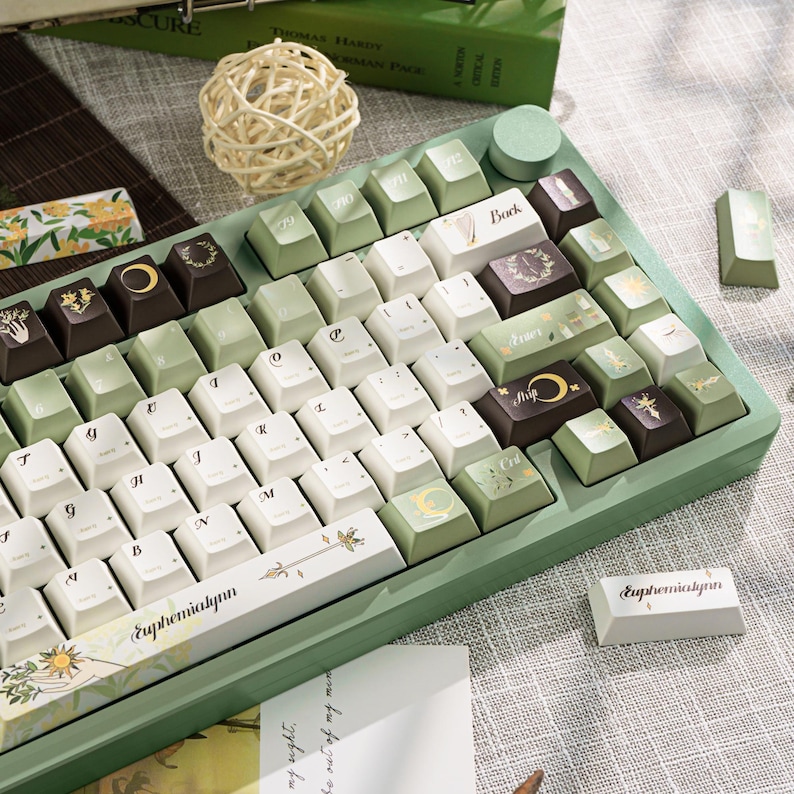Puede incluir: Un teclado mec&aacute;nico personalizado con un marco verde claro y teclas en blanco, verde claro y marr&oacute;n. Las teclas presentan varios dise&ntilde;os, incluyendo patrones florales, lunas y texto. El teclado tiene una est&eacute;tica &uacute;nica.