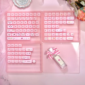 May include: A pink keyboard with white keycaps featuring cartoon cat designs. The keyboard is divided into four sections, with a perfume bottle and a pink and white striped bow in the bottom right corner. The keycaps have letters and numbers.