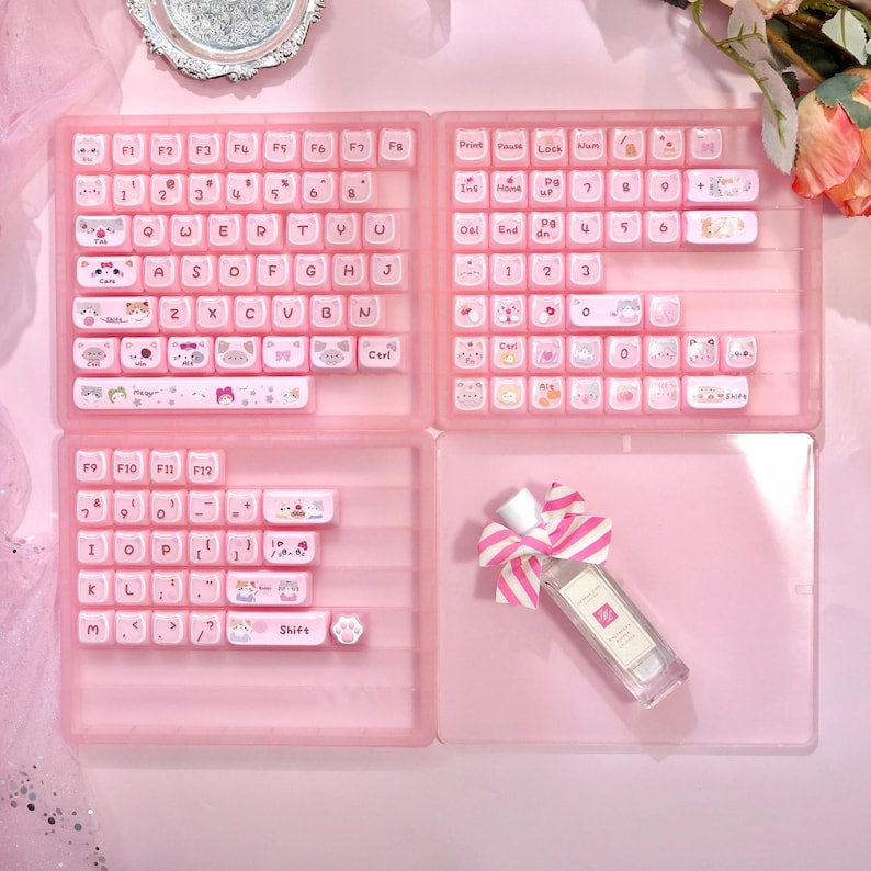 May include: A pink keyboard with white keycaps featuring cartoon cat designs. The keyboard is divided into four sections, with a perfume bottle and a pink and white striped bow in the bottom right corner. The keycaps have letters and numbers.