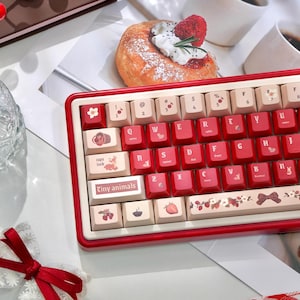 Può includere: Una tastiera meccanica rossa e crema con tasti personalizzati. I tasti presentano varie illustrazioni, tra cui un cervo, una fragola e un fiocco. La tastiera ha il testo "Tiny animals" sulla barra spaziatrice.